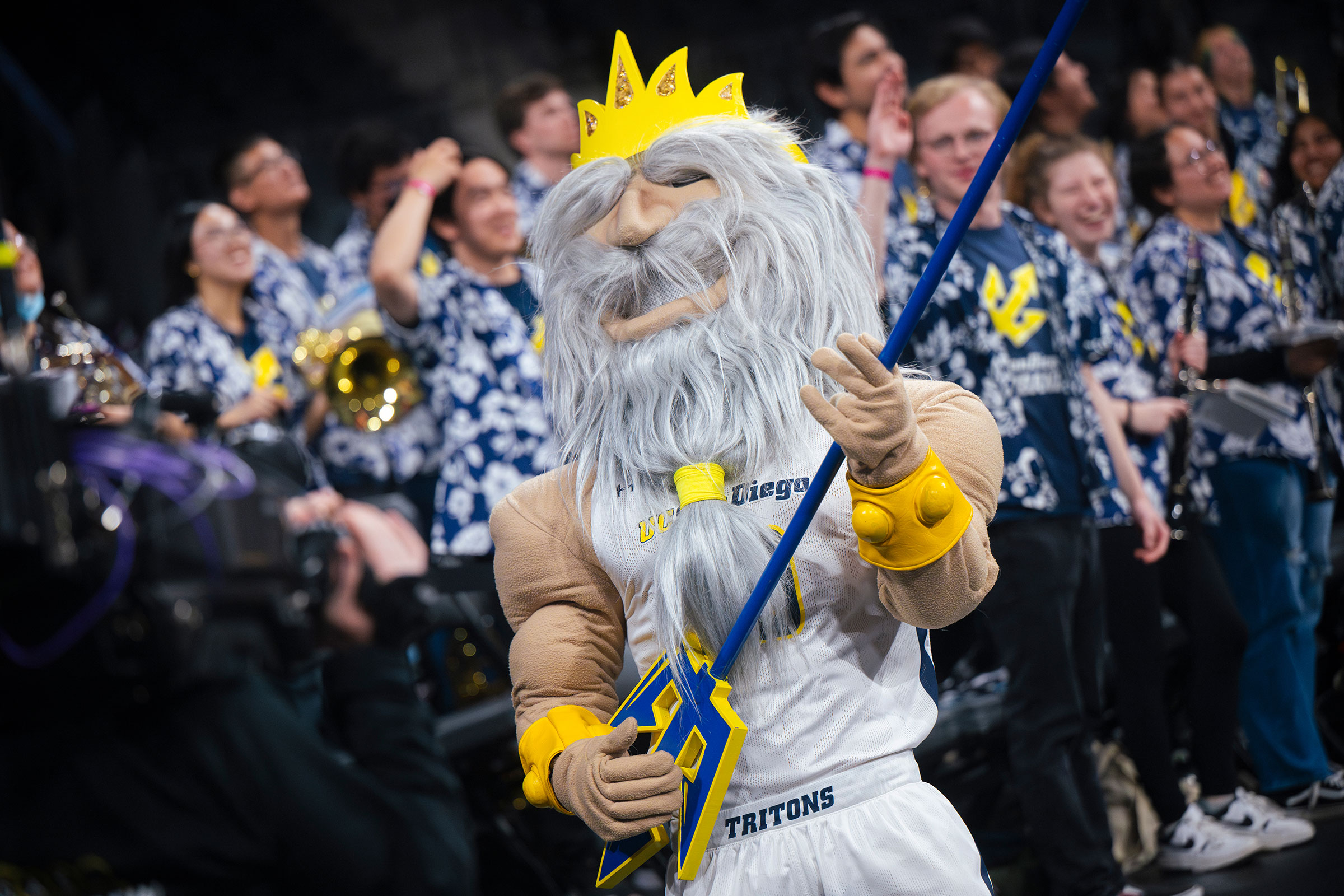 A King Triton mascot dressed in a basketball uniform pretends to play a trident as a guitar.